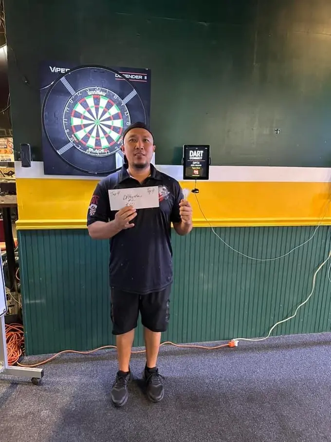 Darts player standing in front of a dartboard and holding a paper certificate
