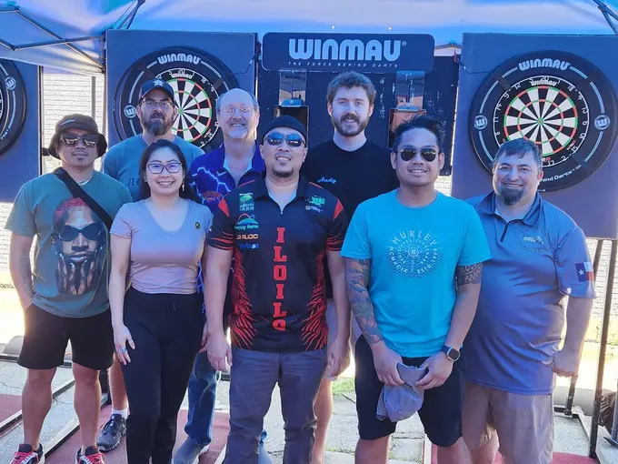 Six darts players smiling together in front of tournament boards