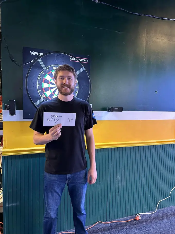 Darts player smiling and holding a tournament note in front of a dartboard