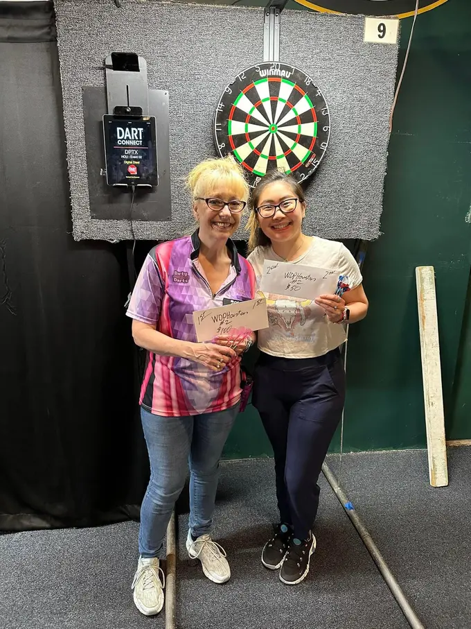 Two women smiling with papers in front of an electronic scoreboard and dartboard