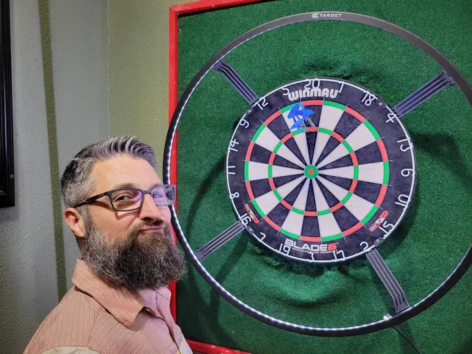Bearded darts player standing beside a round dartboard with red surround