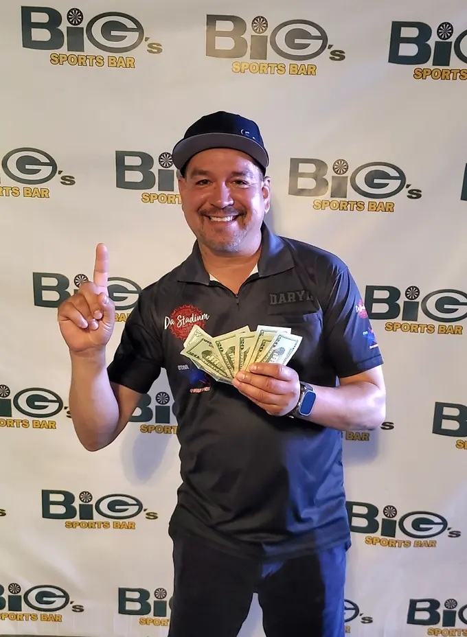 Smiling darts competitor holding prizes in front of a Big's Sports Bar sponsor backdrop
