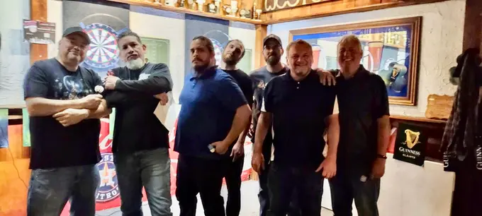Six darts players posing together indoors with a dartboard in the background
