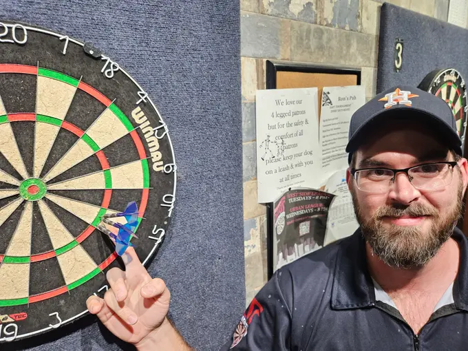 Darts player in a cap smiling and gesturing beside a steel-tip dartboard
